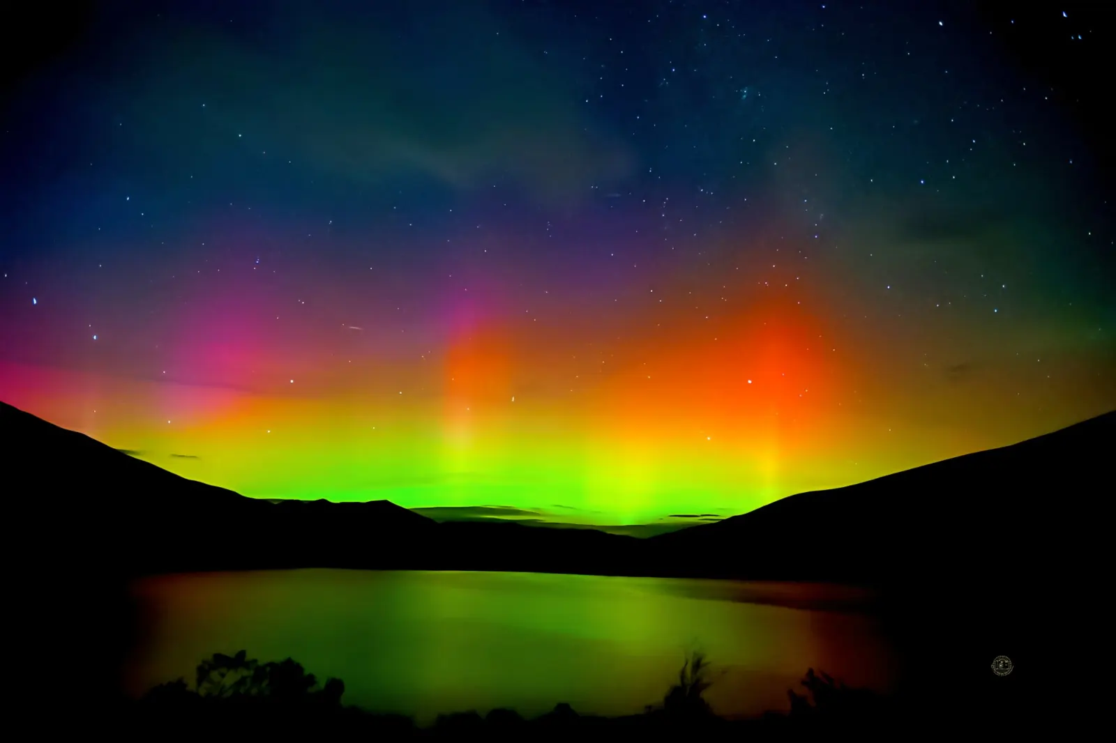 Aurora borealis in green, orange and magenta over silhouetted hills, reflected in still water